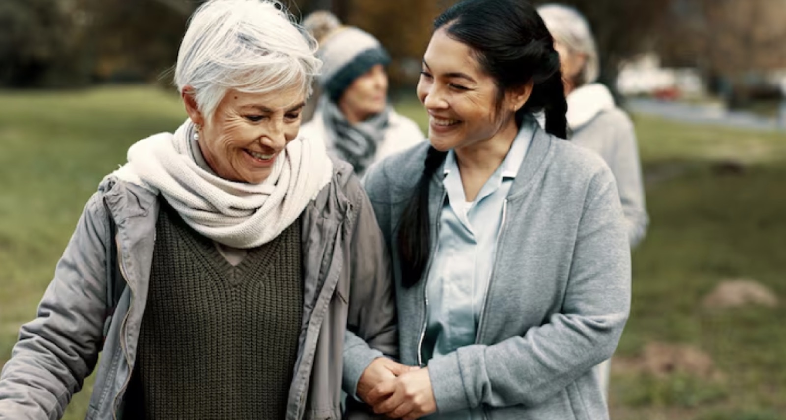 A carer walks with an older woman in a park.