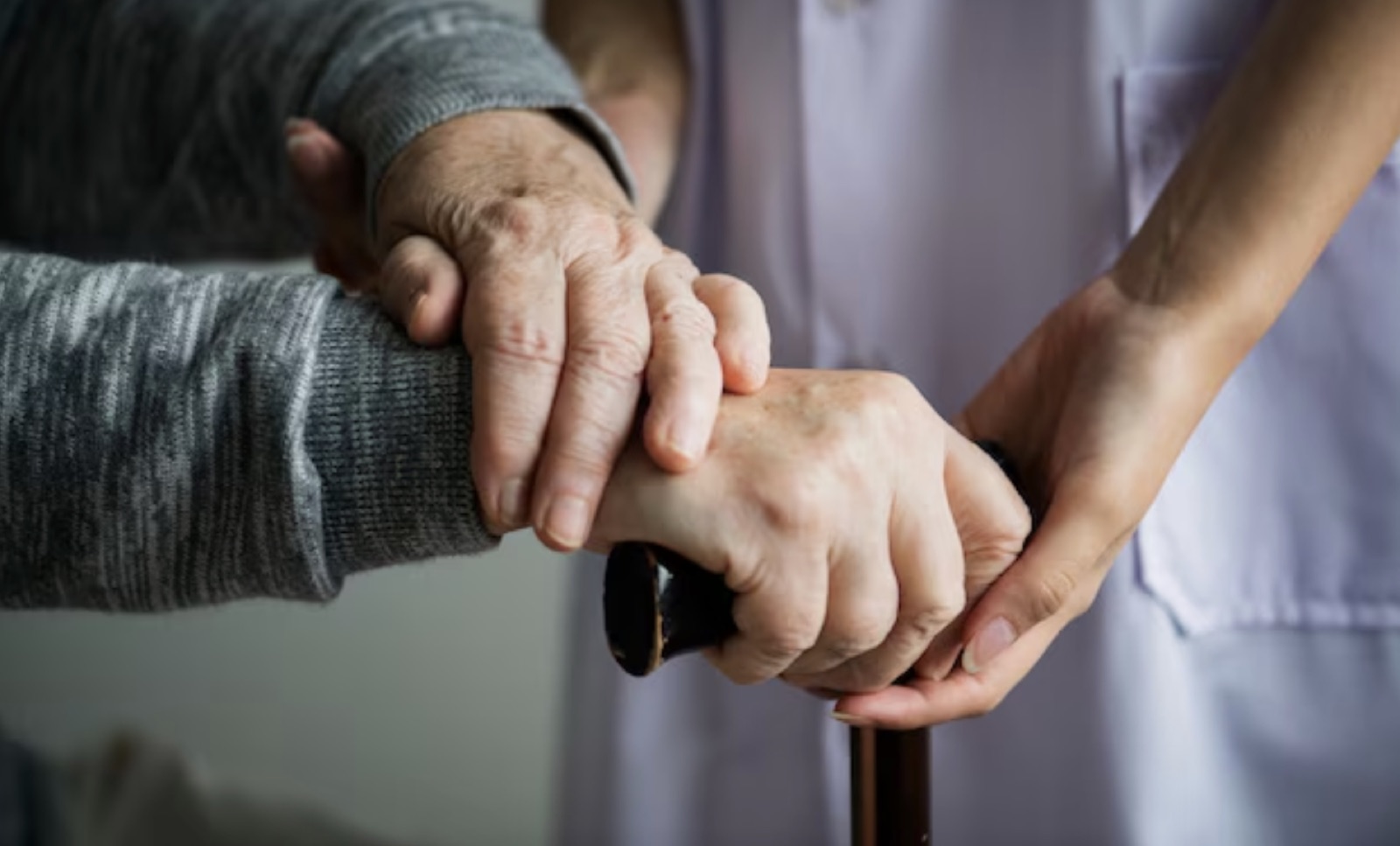 Aged hands on a walking cane with a carer's hand offering support.
