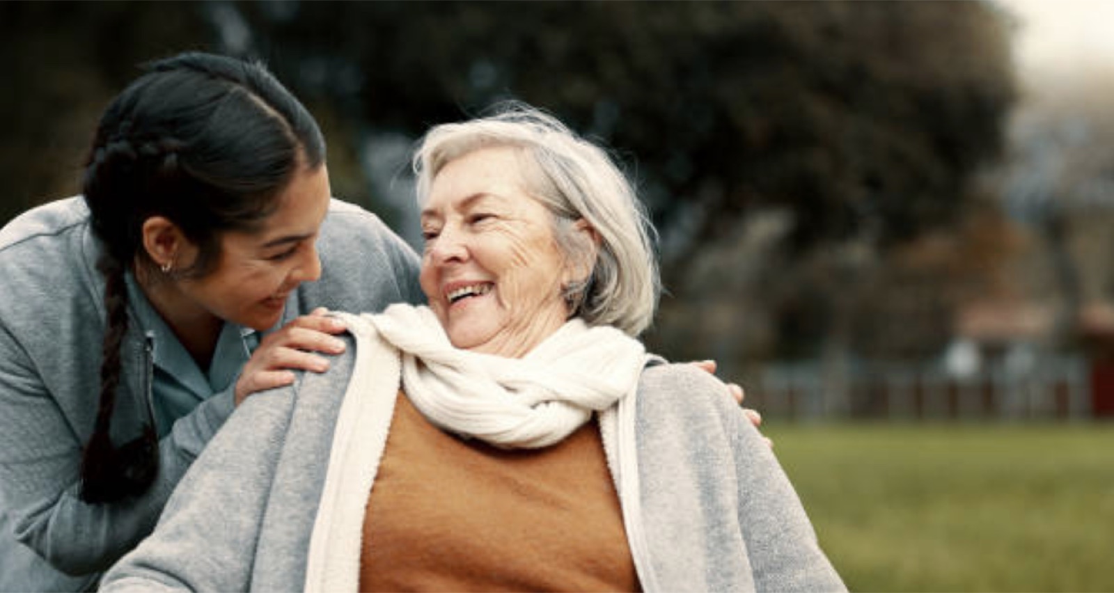A care worker and an older woman share a moment of laughter on a sunny garden bench — a glimpse of everyday Enrich Home Supports care.